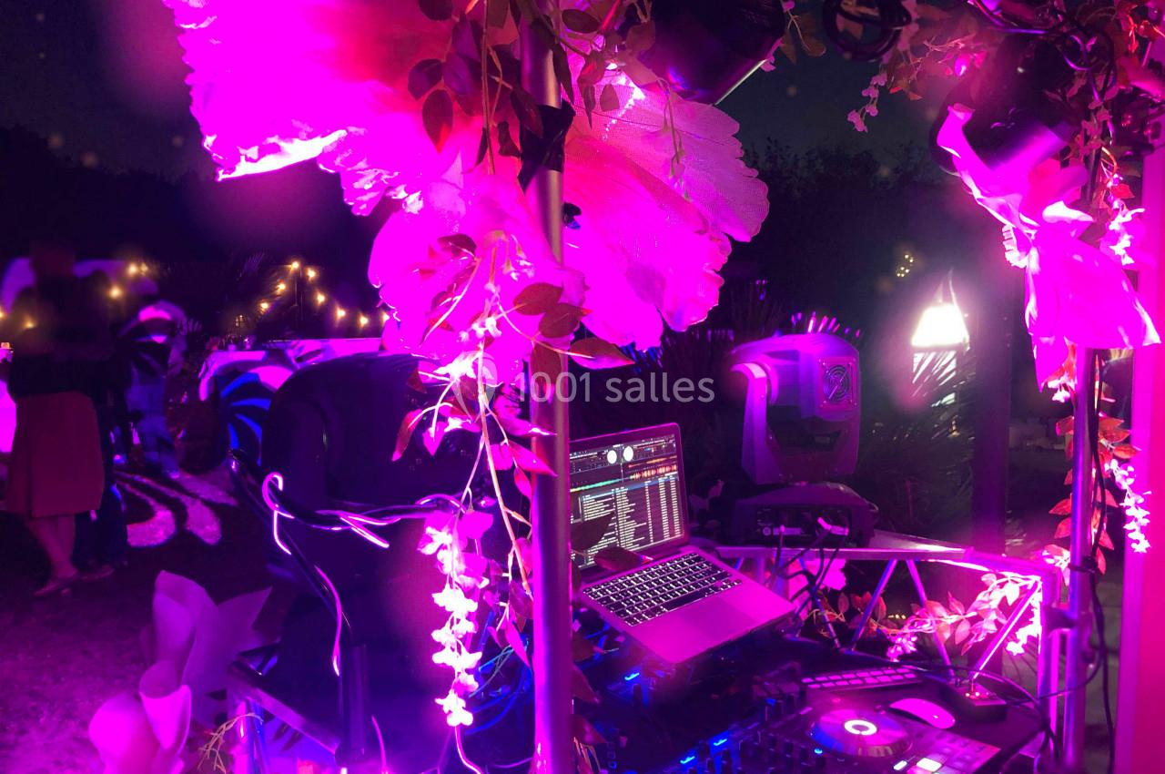 Poste de DJ en extérieur, éclairé par des lumières roses et entouré de décorations florales, avec des invités en arrière…