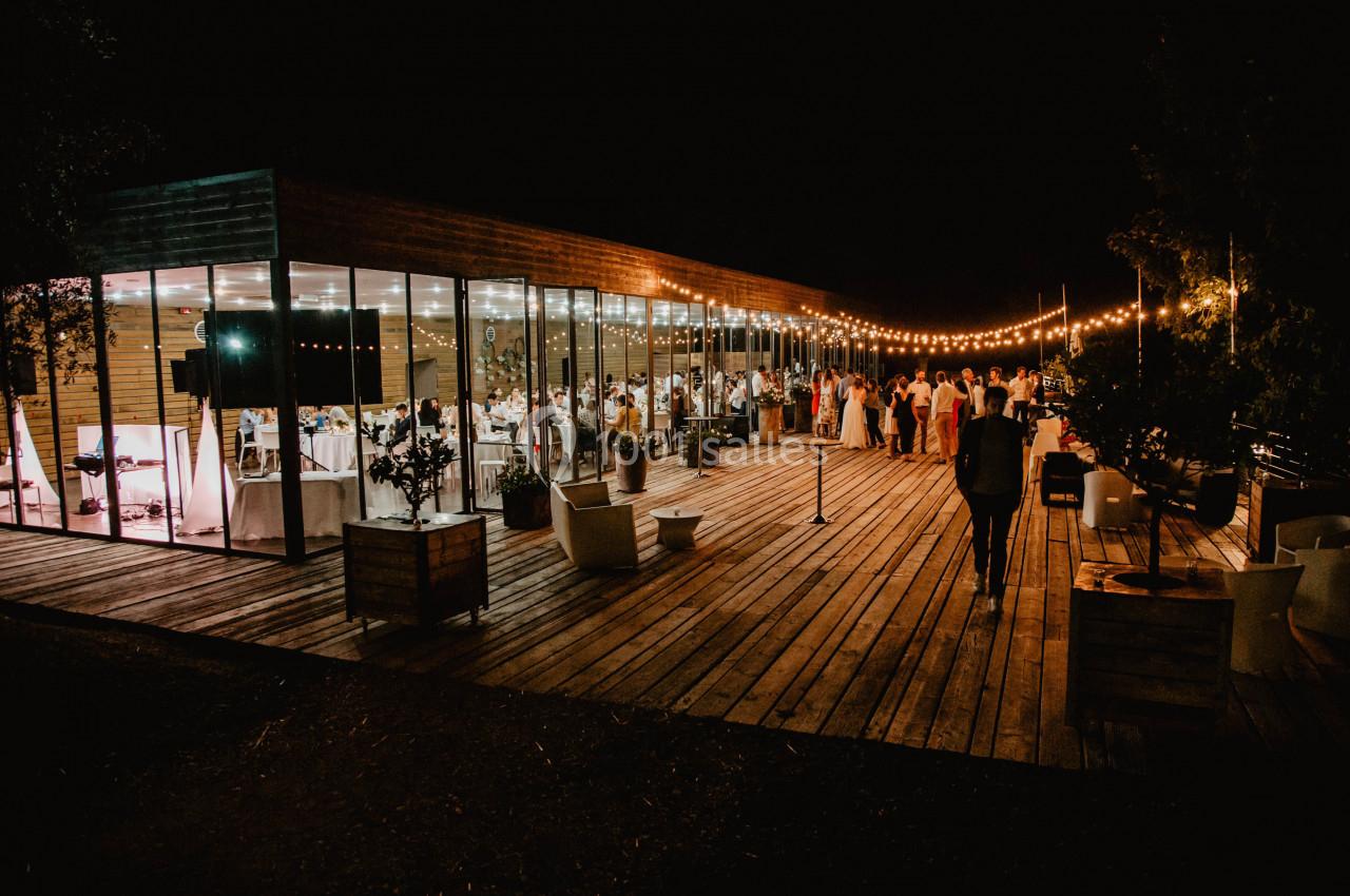Terrasse en bois éclairée par des guirlandes lumineuses, avec des invités rassemblés près d'un bâtiment vitré.