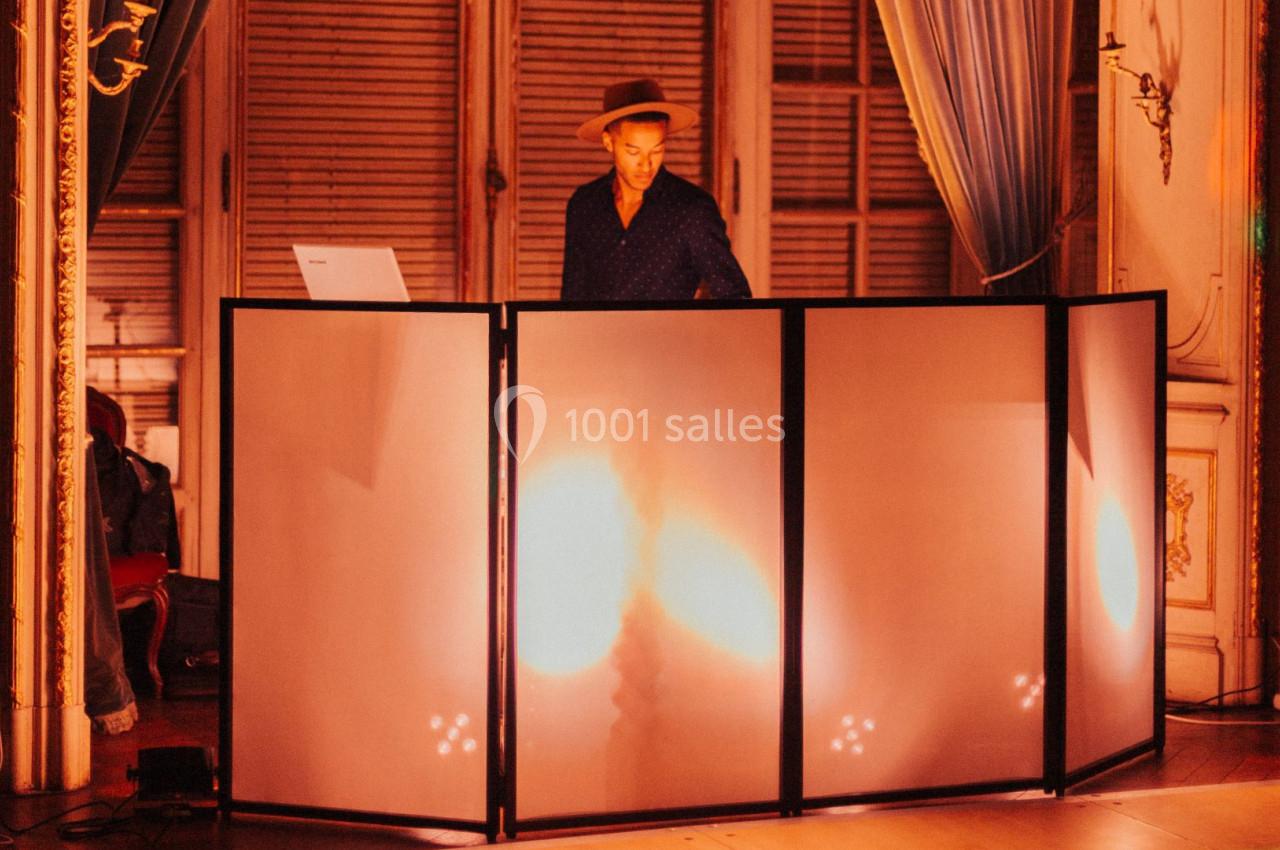 Un DJ mixe derrière une table éclairée dans une salle élégante avec rideaux et boiseries.