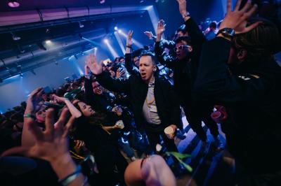 Des personnes dansent et lèvent les bras dans une ambiance festive, éclairée par des lumières colorées.