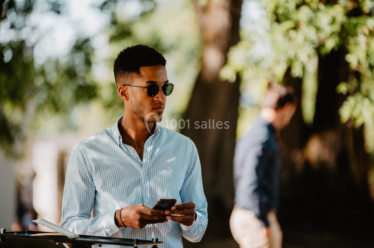Un homme portant des lunettes de soleil utilise son téléphone dans un parc, avec des arbres en arrière-plan.