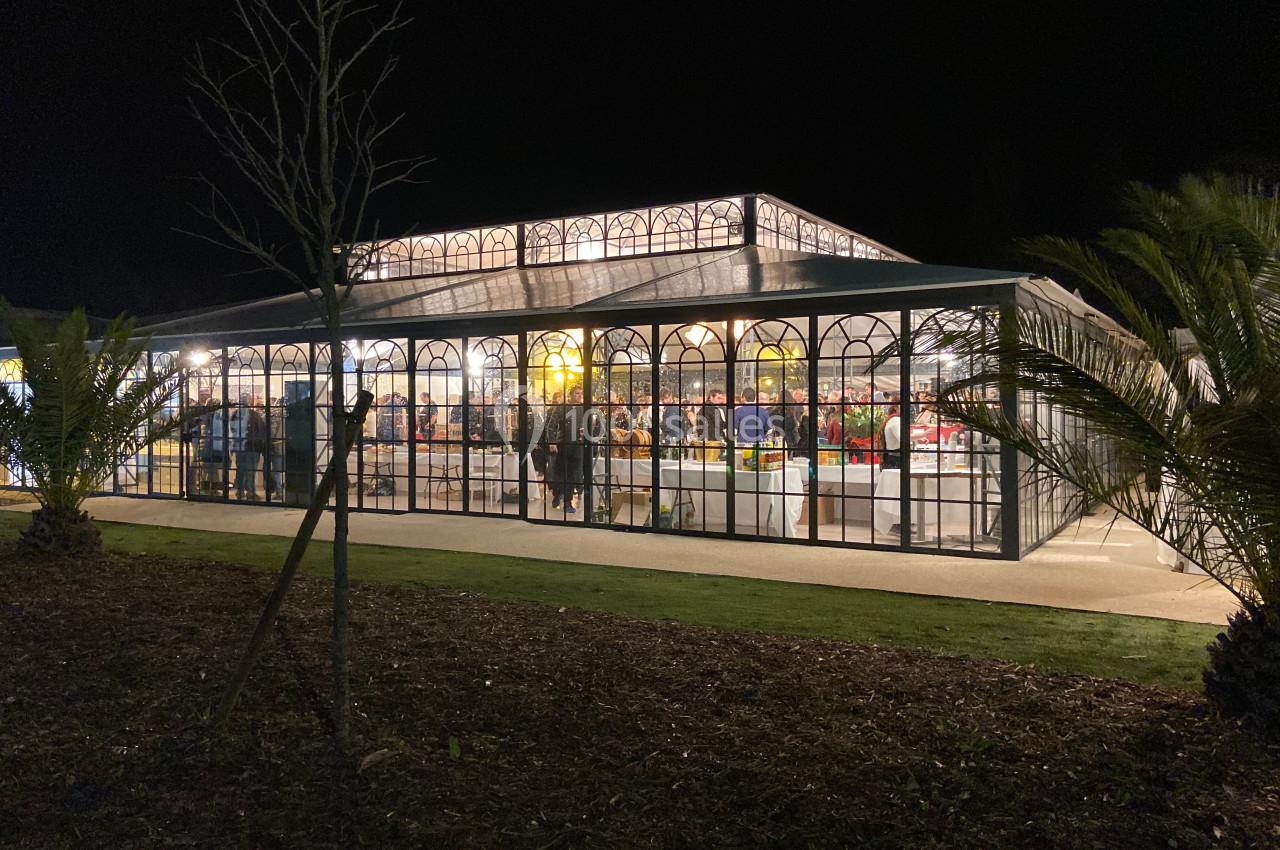 Vue nocturne d'une serre illuminée avec des vitrines en verre, abritant des stands et des visiteurs à l'intérieur.