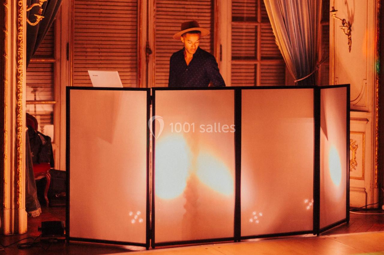 Un DJ mixe derrière une table éclairée dans une salle ornée de rideaux et de boiseries.