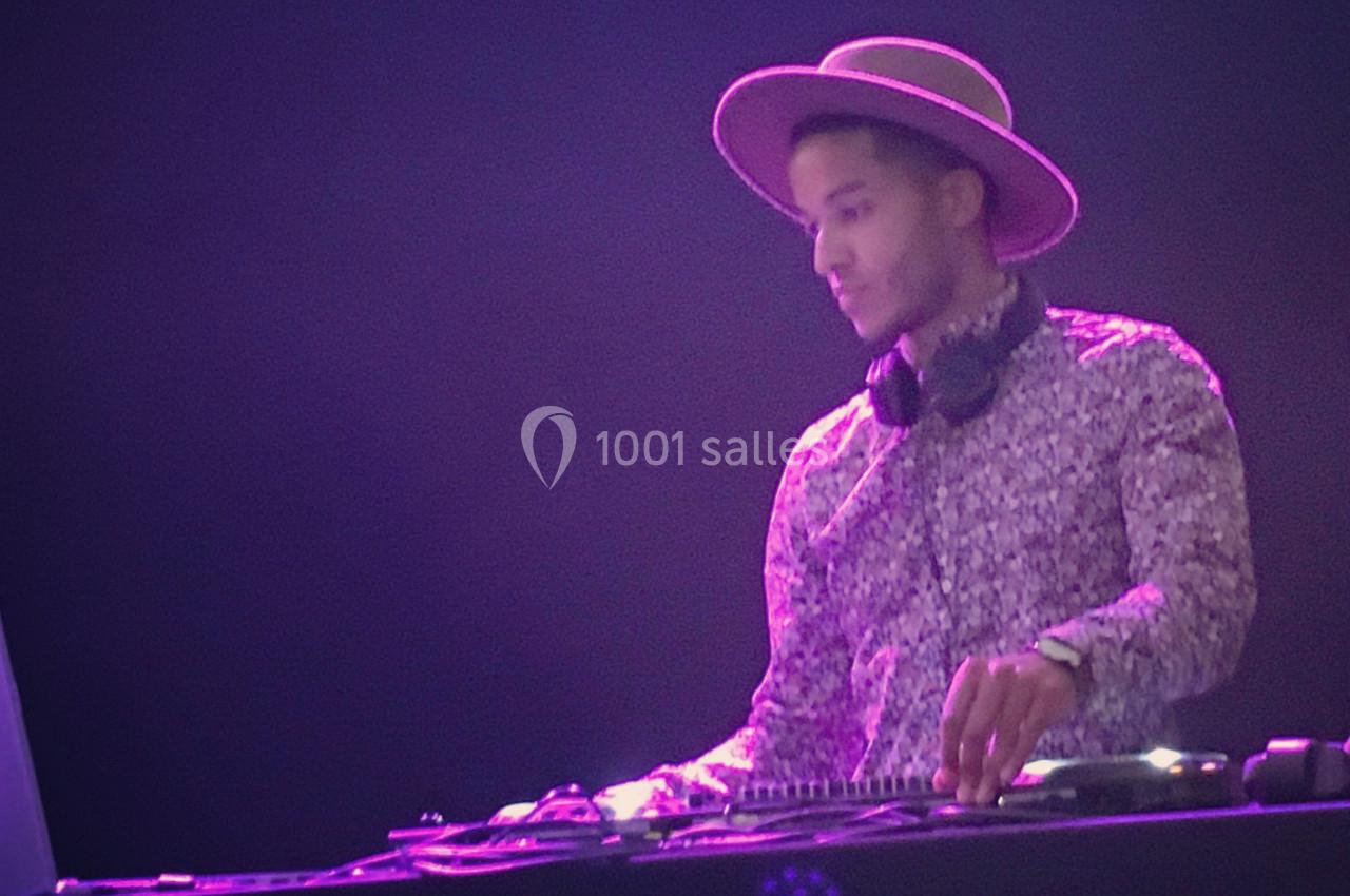 Un DJ portant un chapeau et un casque autour du cou mixe de la musique sur une table de mixage dans une ambiance lumineuse.