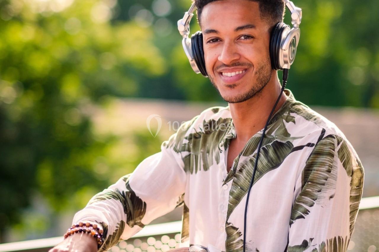 Un DJ souriant mixe de la musique en extérieur, entouré de verdure, avec un ordinateur portable à ses côtés.