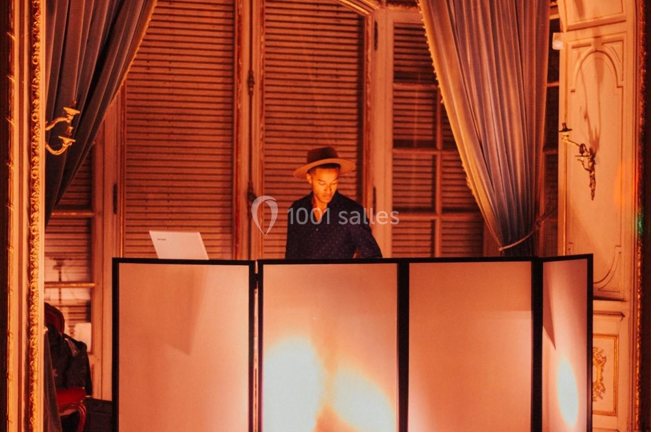 Un DJ mixe derrière une table éclairée dans une salle élégante avec rideaux et moulures.