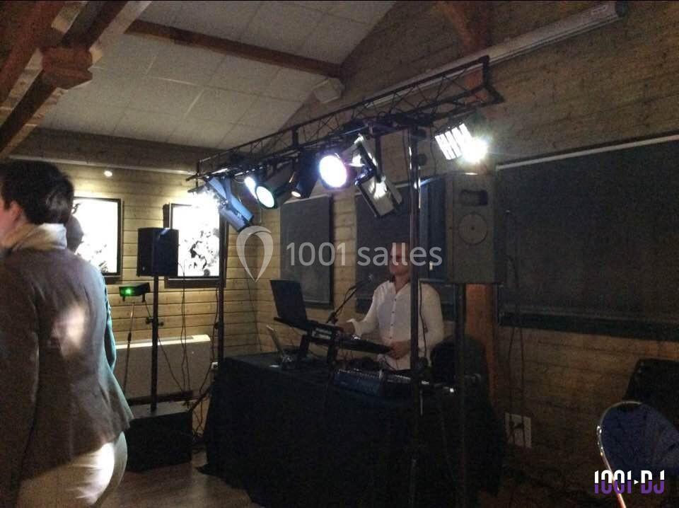 Un DJ joue de la musique sur une scène éclairée dans une salle en bois, avec des personnes dansant au premier plan.