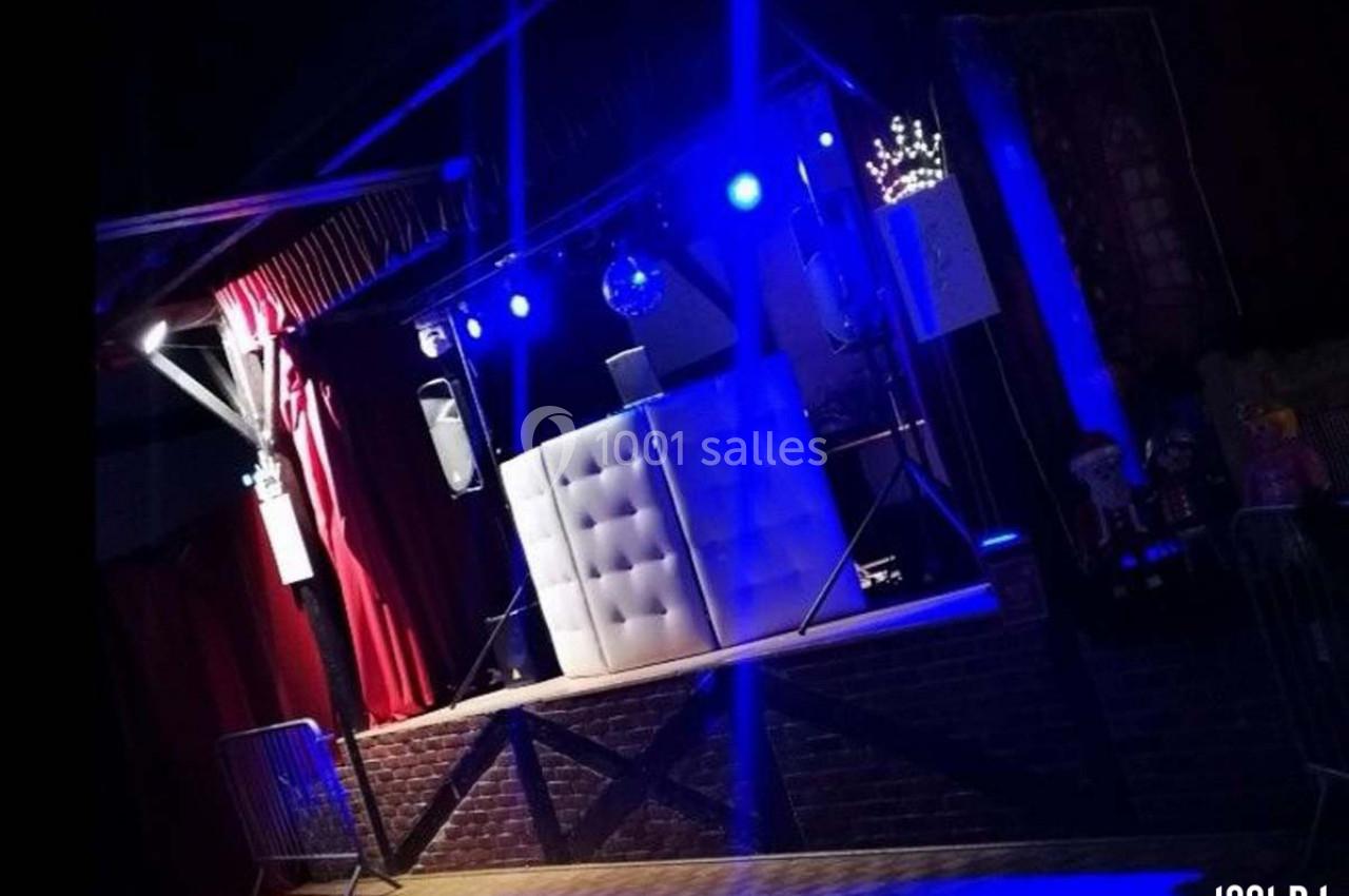 Scène éclairée en bleu avec une table de DJ blanche, des rideaux rouges et des équipements audio.