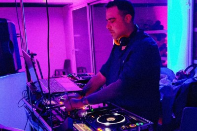 Un DJ en chemise blanche utilise une table de mixage avec des écrans d'ordinateur en arrière-plan.