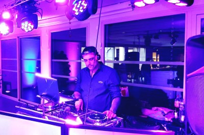 Un DJ en chemise blanche utilise une table de mixage avec des écrans d'ordinateur en arrière-plan.