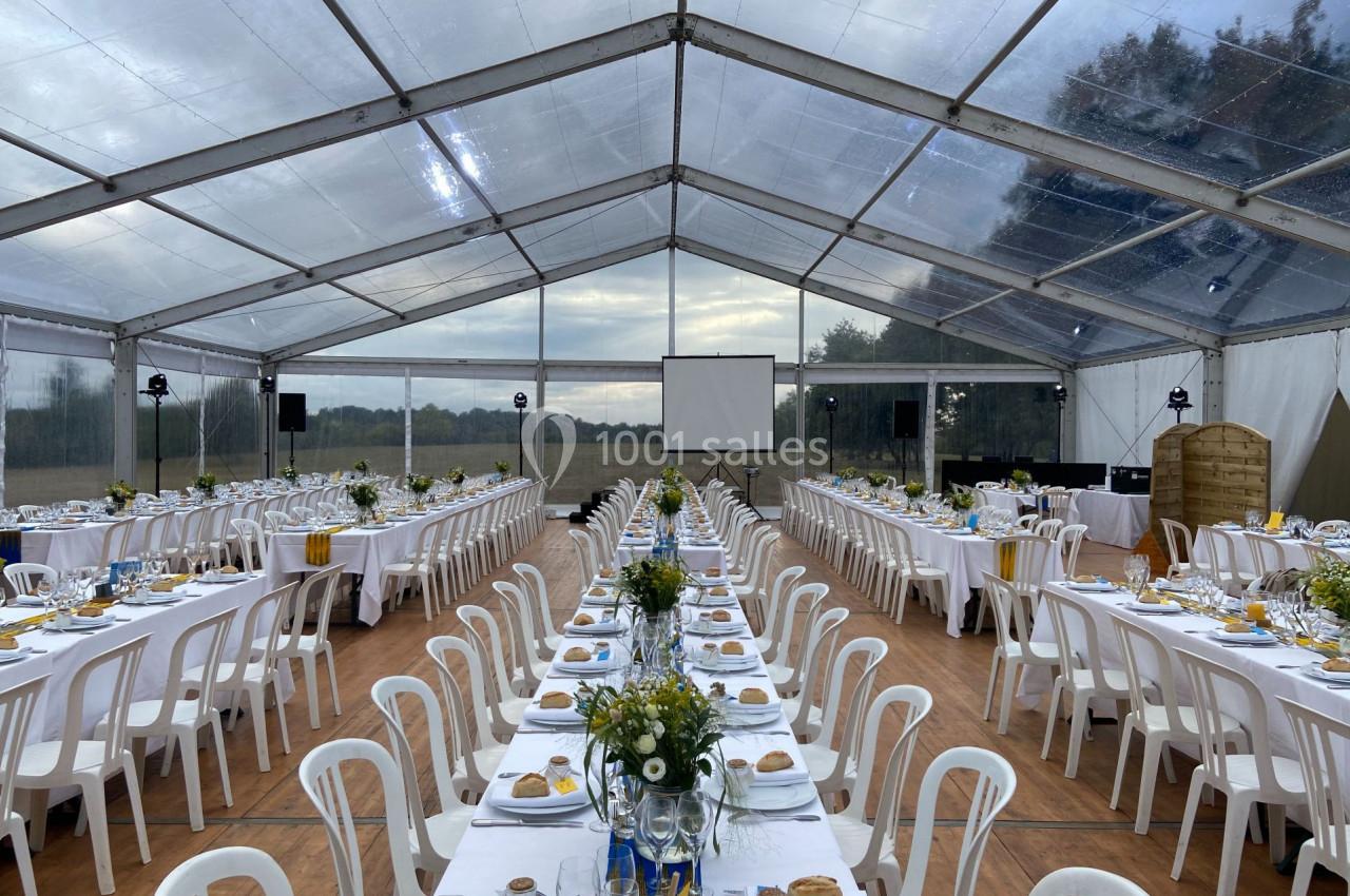 Grande tente transparente aménagée avec des tables alignées, décorées de fleurs et de vaisselle pour un événement.