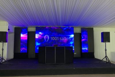 Groupe de musiciens jouant sur une scène couverte en plein air, éclairée par des lumières colorées, avec un public devant.