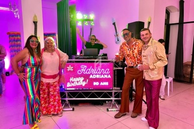 Un DJ souriant anime une soirée dansante dans une salle éclairée par des lumières colorées, avec des invités sur la piste.