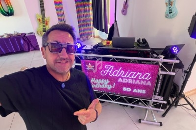 Un DJ souriant anime une soirée dansante dans une salle éclairée par des lumières colorées, avec des invités sur la piste.