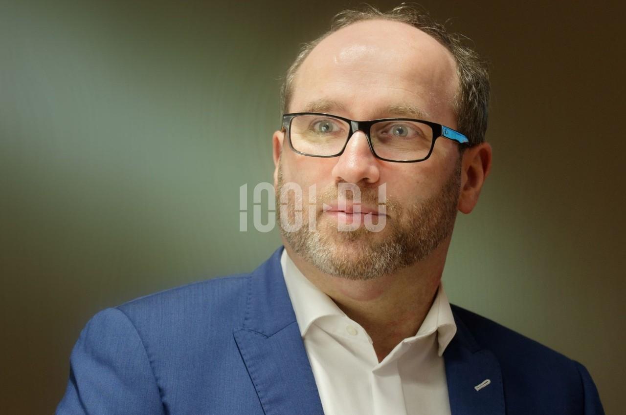 Homme portant des lunettes et un costume bleu, regardant légèrement sur le côté dans un environnement intérieur.