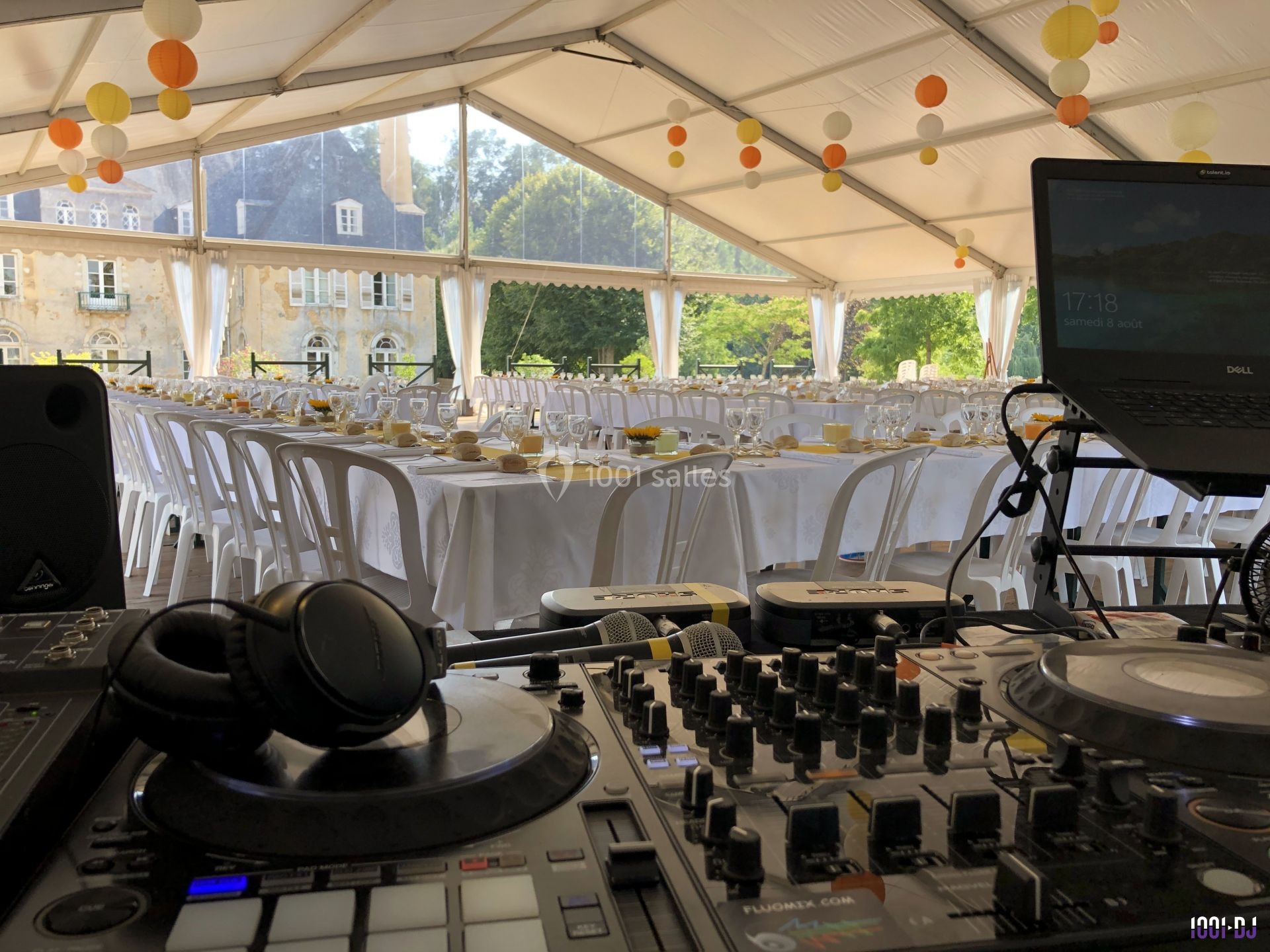 Tables dressées pour un événement sous une tente, avec une console DJ au premier plan et un bâtiment en arrière-plan.