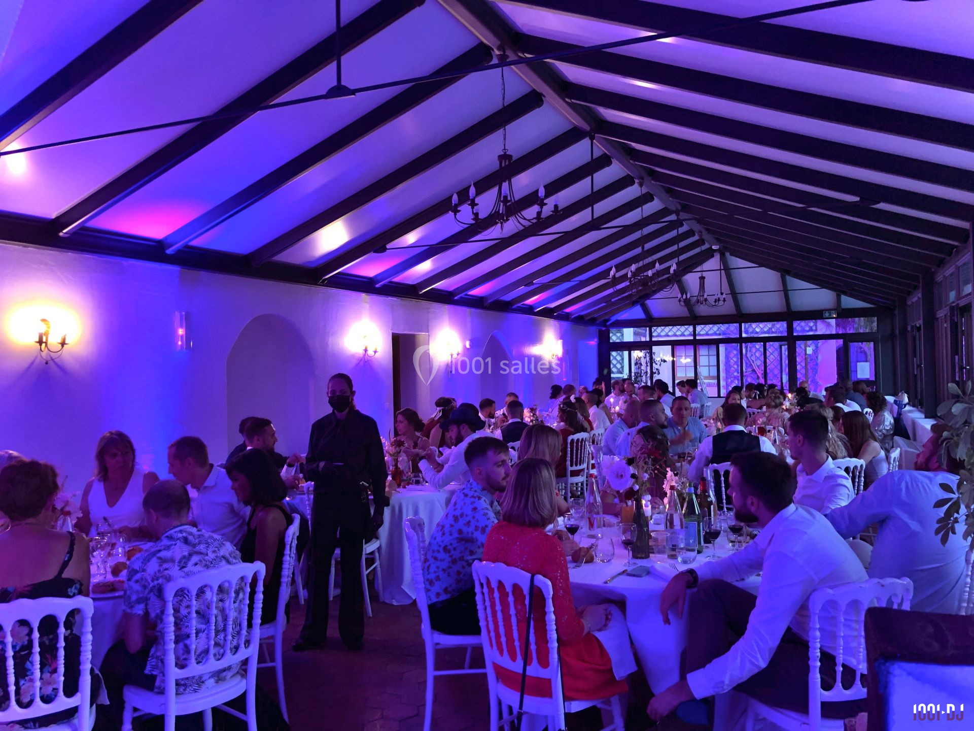 Salle de réception éclairée en violet, remplie de convives assis à des tables rondes lors d'un événement festif.