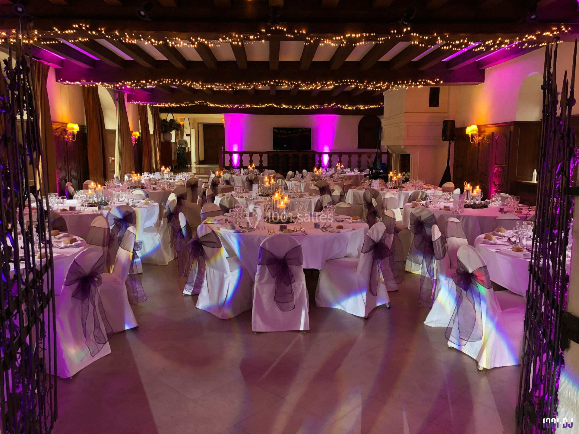 Salle de réception décorée pour un événement, avec tables dressées, chaises ornées de rubans et guirlandes lumineuses au…