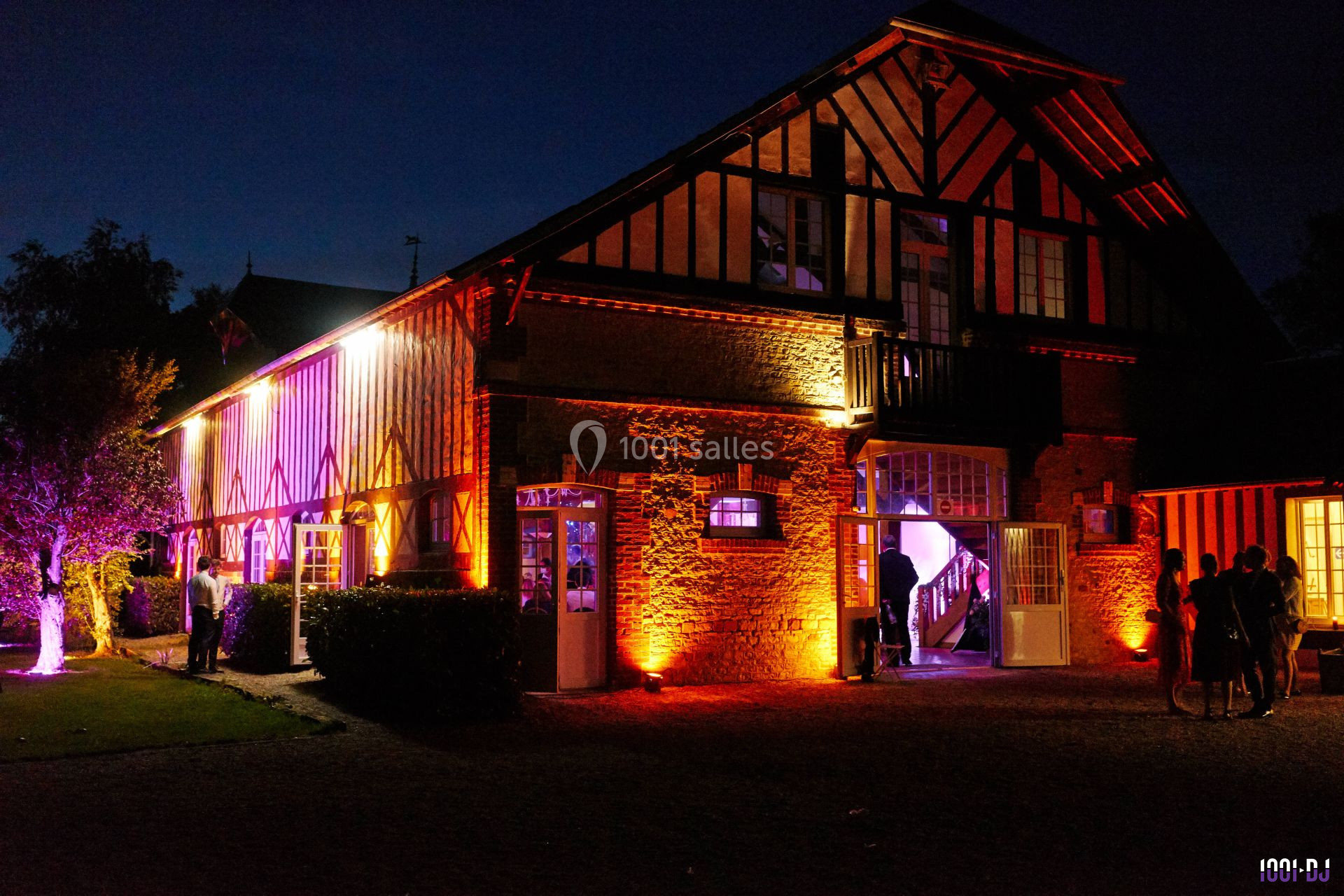 Façade d'une grande maison à colombages éclairée par des lumières colorées en soirée, avec quelques personnes à l'extérieur.