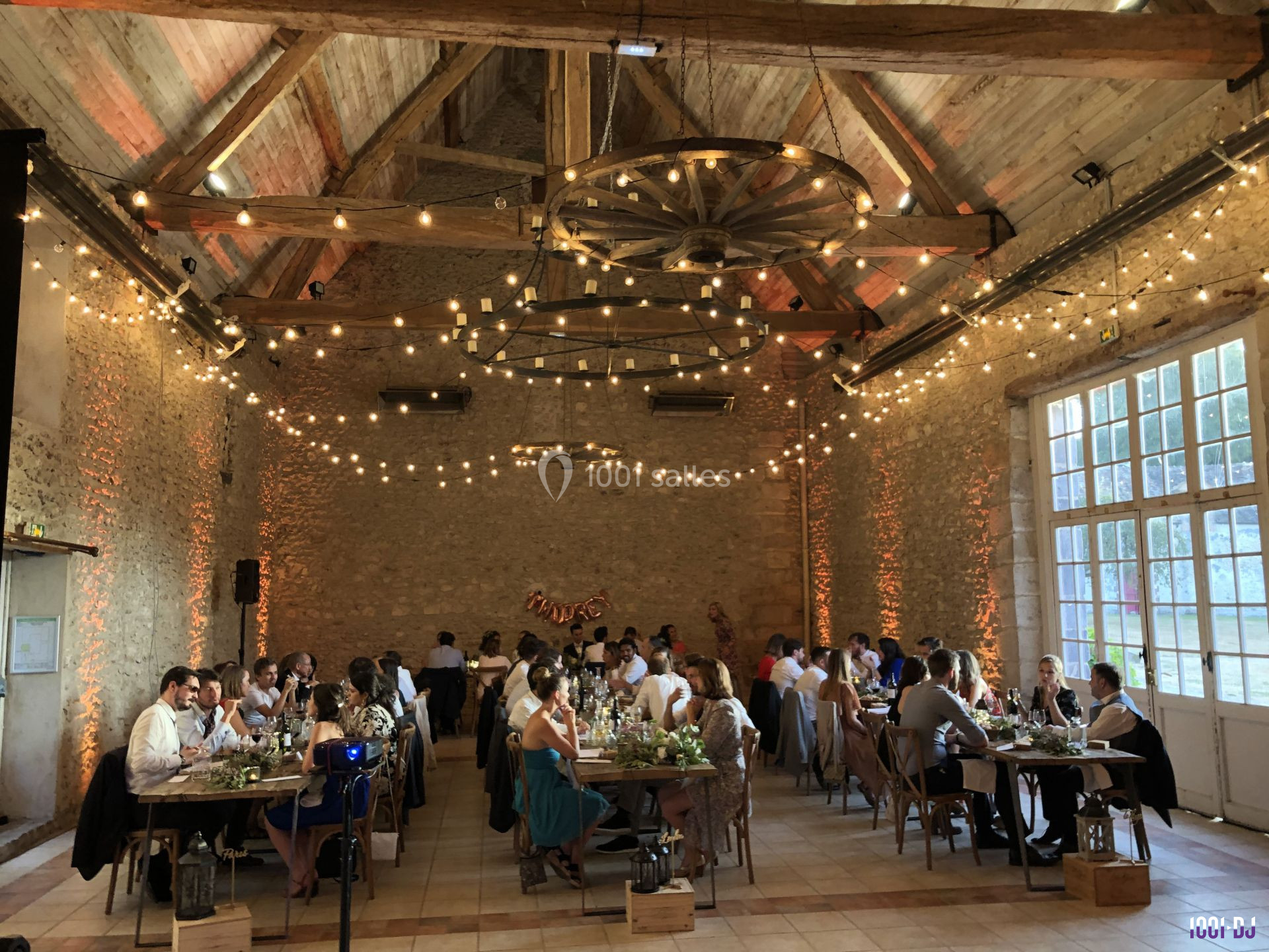 Salle de réception en pierre avec des invités dînant à des tables décorées, éclairée par des guirlandes lumineuses.