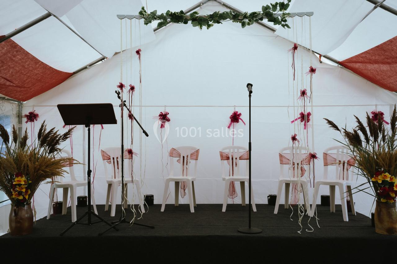 Estrade décorée pour une cérémonie avec chaises blanches ornées de rubans roses, micros et arrangements floraux.