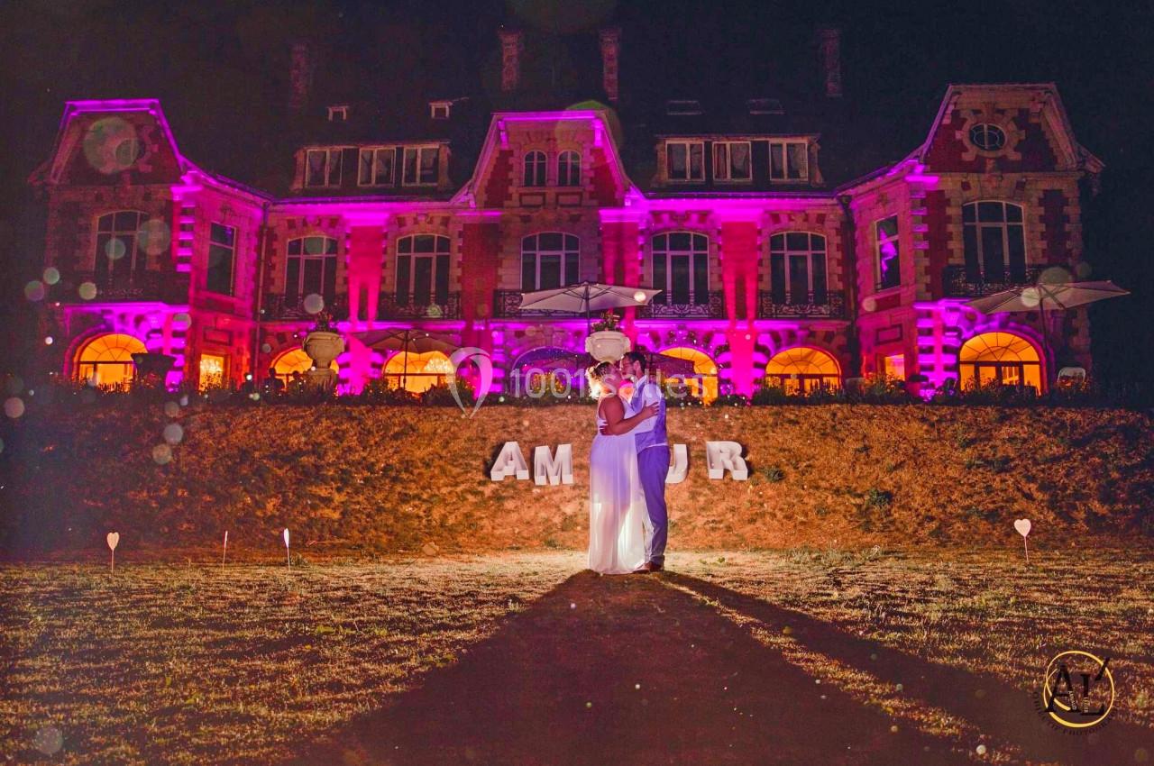 Un couple s'embrasse de nuit devant un bâtiment illuminé en rose, avec le mot ’AMOUR’ en lettres blanches au sol.
