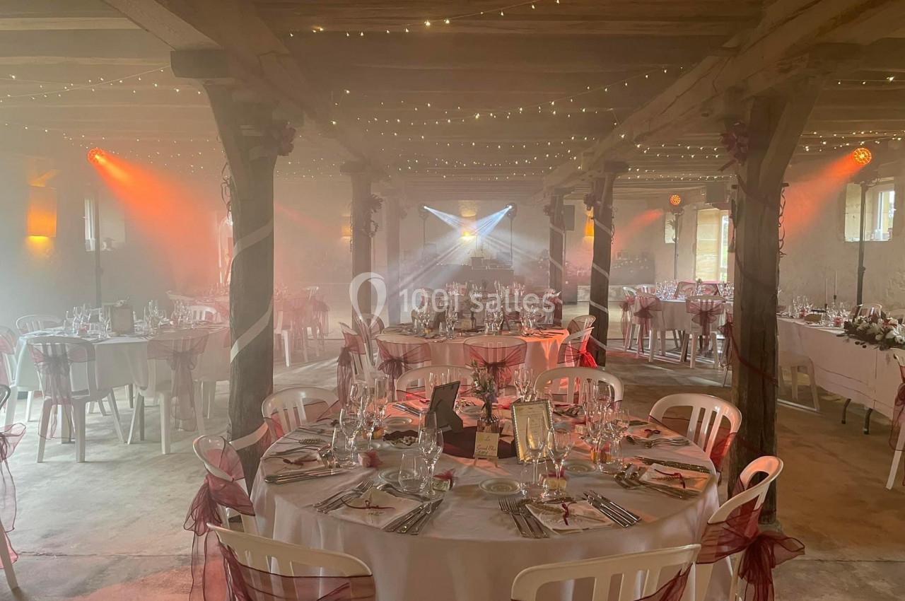 Salle de réception décorée avec des tables rondes, nappes blanches, chaises ornées de rubans rouges et éclairage tamisé.
