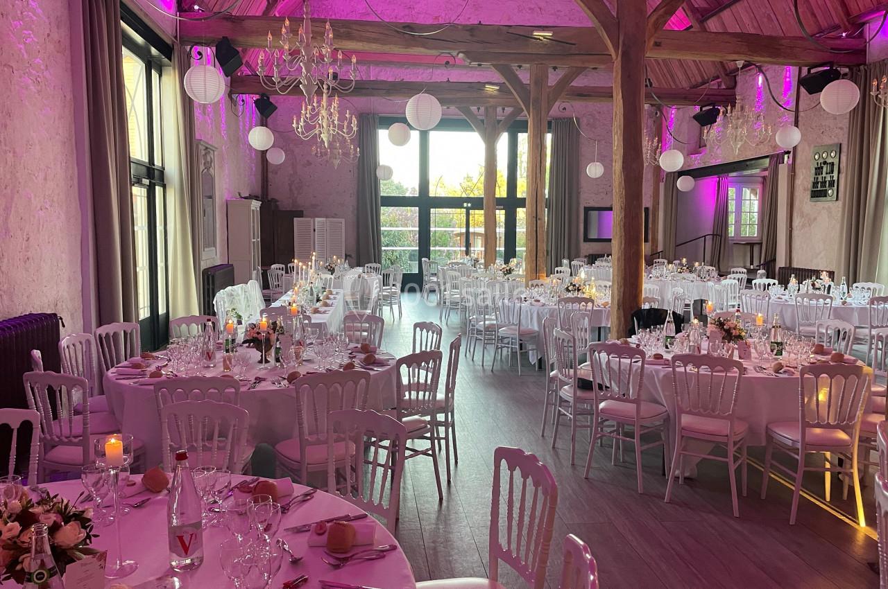Salle de réception décorée pour un mariage, avec tables rondes dressées, chaises blanches et éclairage rose.