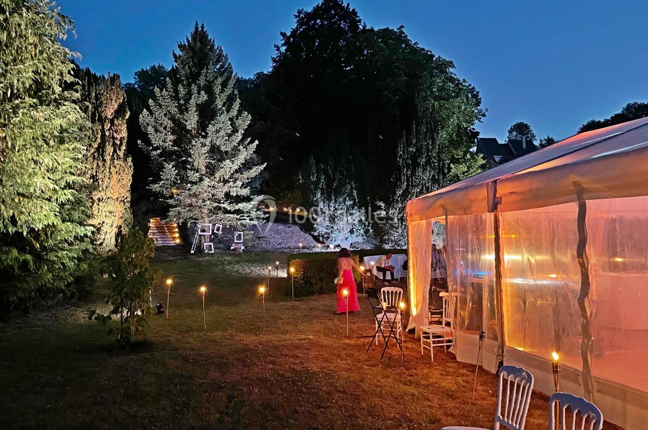 Jardin éclairé en soirée avec une tente transparente, des chaises blanches et des torches disposées sur la pelouse.