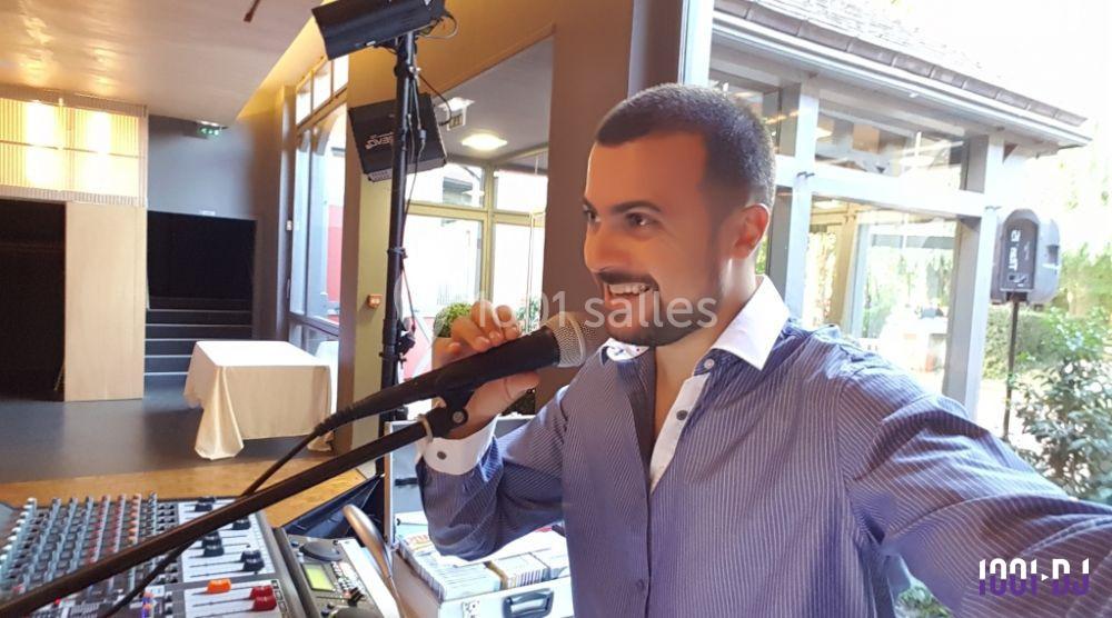 Homme souriant parlant dans un micro, debout près d'une table de mixage dans une salle lumineuse.