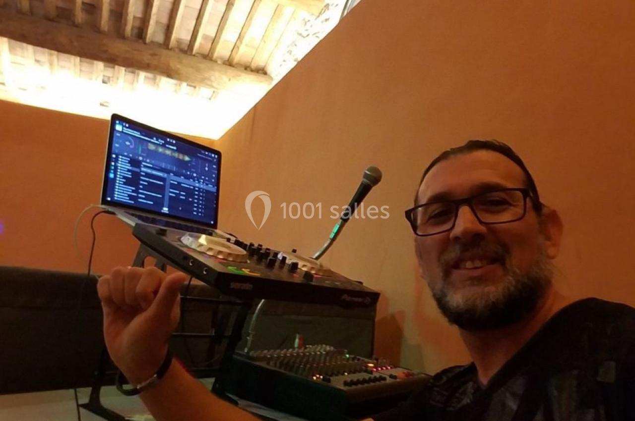 Homme souriant devant un équipement de DJ avec ordinateur, console de mixage et microphone dans une pièce au plafond en bois.