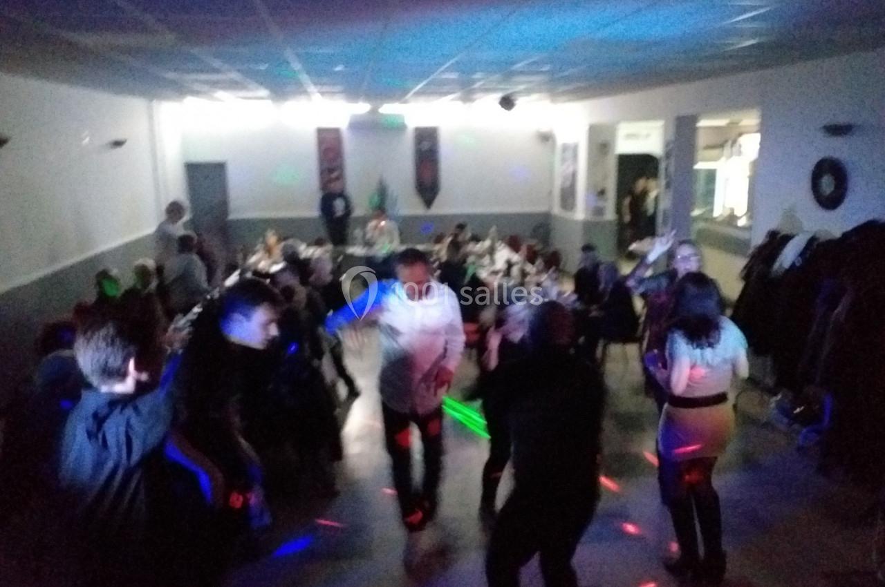 Des personnes dansent dans une salle éclairée par des lumières colorées, avec des tables et des invités en arrière-plan.