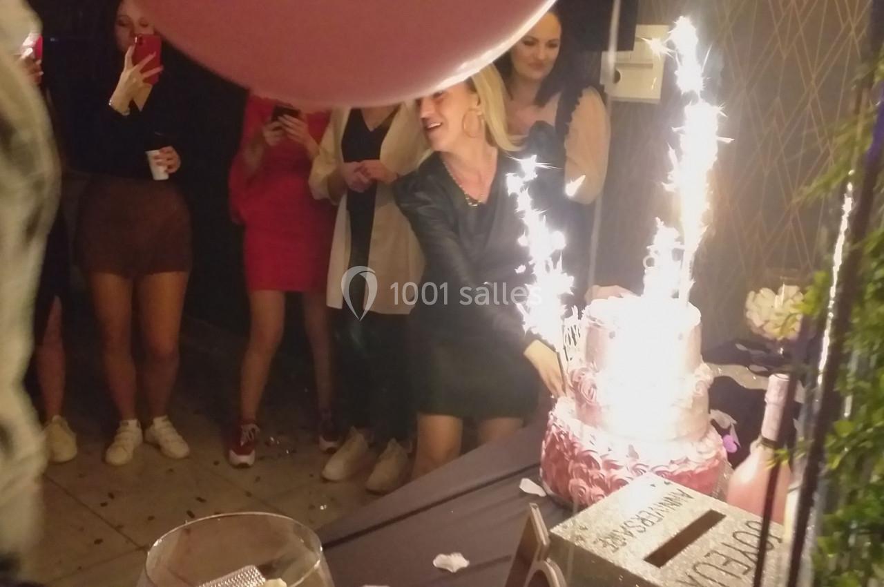 Une femme souriante coupe un gâteau décoré de bougies étincelantes lors d'une fête, entourée de personnes.