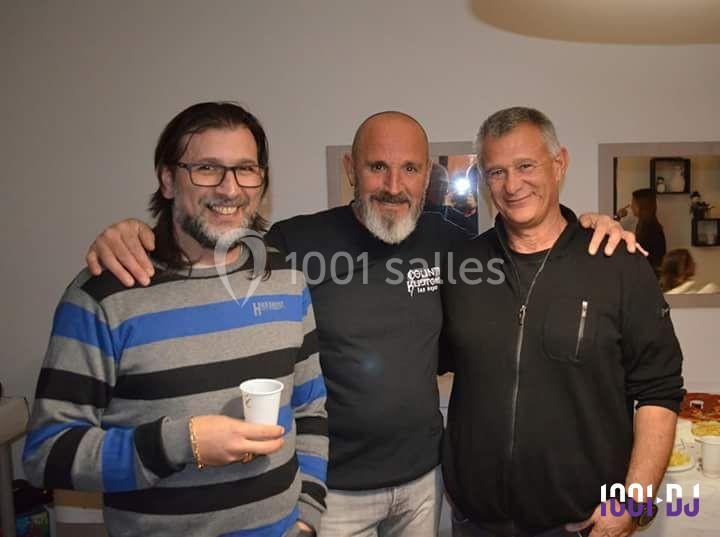 Trois hommes souriants posent ensemble dans une pièce éclairée, l'un tenant un gobelet blanc.