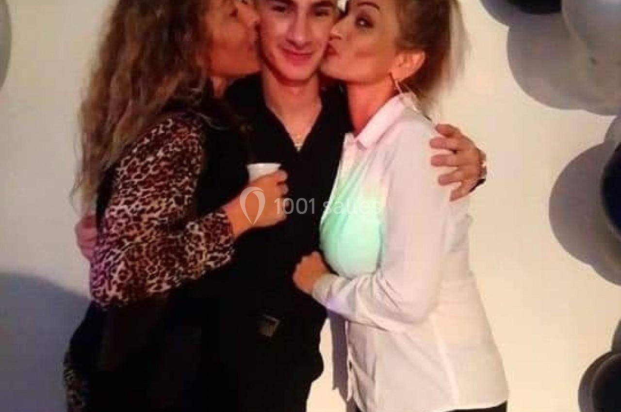 Deux femmes embrassent un jeune homme souriant, debout devant une décoration de ballons noirs, blancs et argentés.