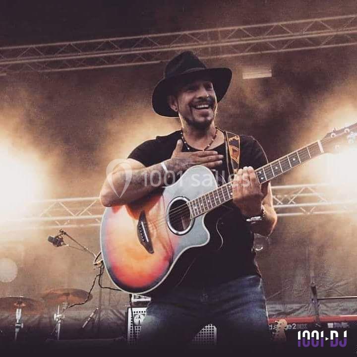 Un musicien souriant joue de la guitare acoustique sur une scène en plein air, entouré de projecteurs et de fumée.