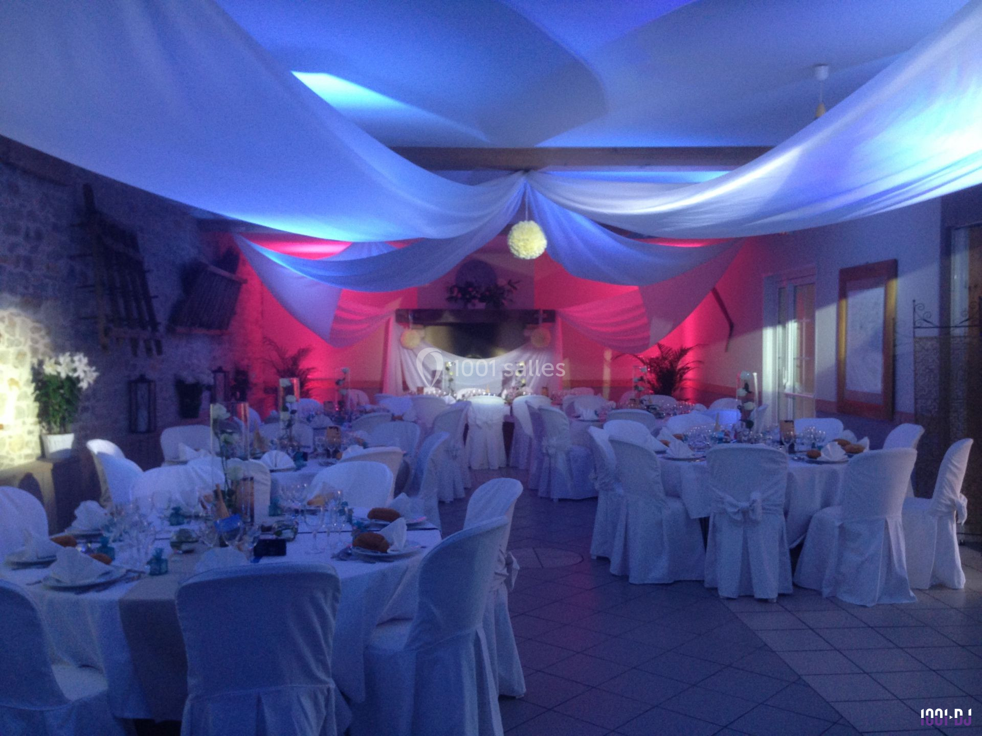 Salle de réception décorée avec des nappes blanches, des éclairages colorés et des drapés suspendus au plafond.