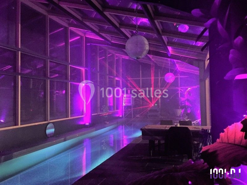 Salle intérieure avec piscine éclairée par des lumières violettes et roses, décorée de plantes et de mobilier en bois.