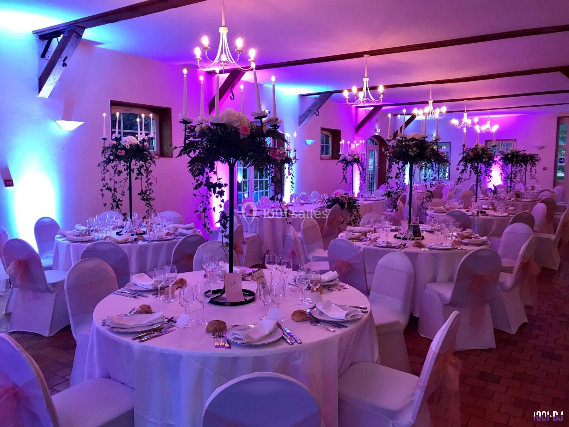 Salle de réception décorée pour un mariage, avec tables rondes dressées, chaises blanches et éclairage rose.