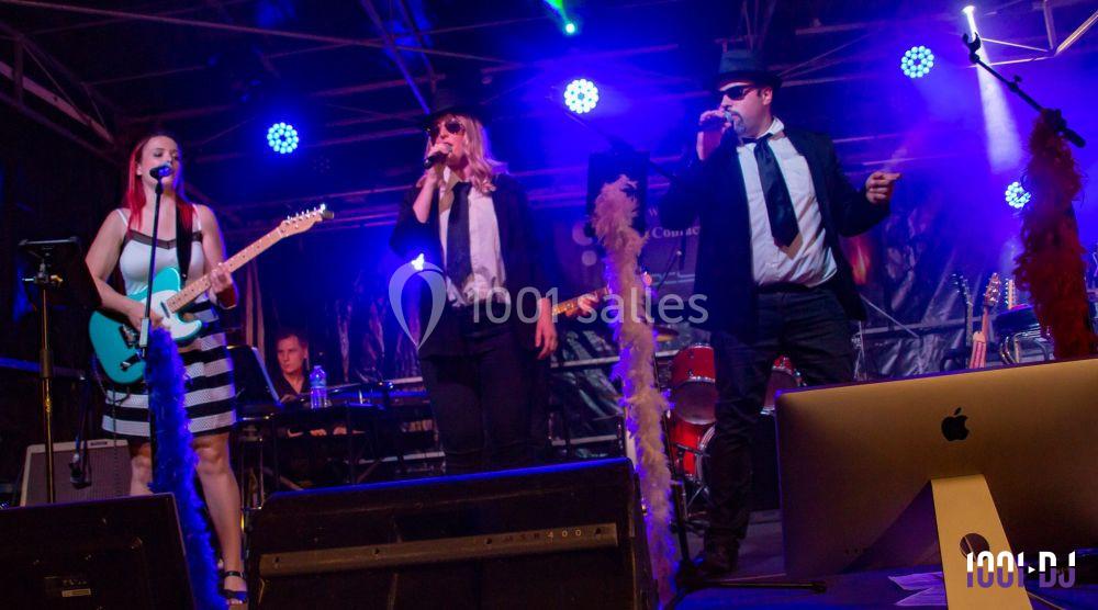 Trois musiciens et une chanteuse sur scène, éclairés par des lumières colorées, jouent devant un public.