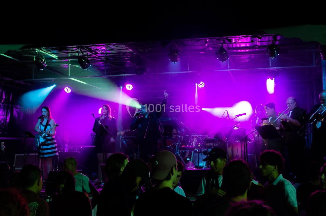 Groupe de musiciens et chanteurs sur scène avec éclairage violet, devant un public en soirée.