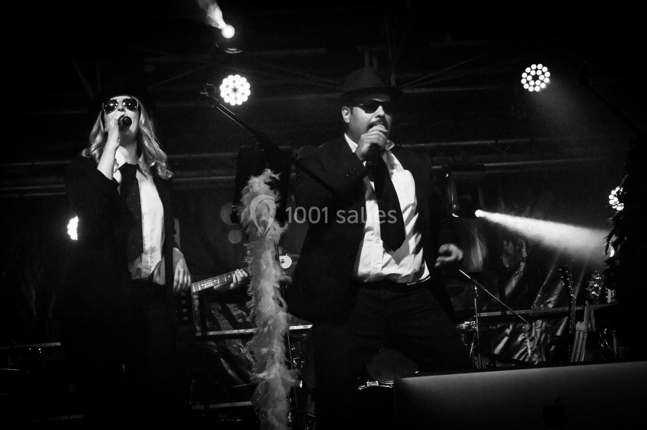 Deux chanteurs en costume sombre et lunettes de soleil se produisent sur une scène éclairée.