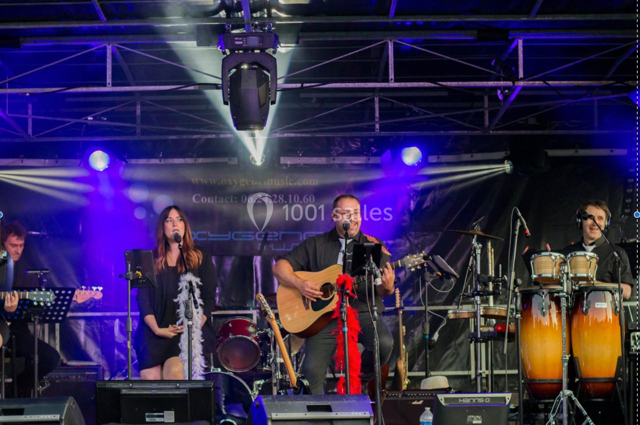 Un groupe de musiciens joue sur une scène éclairée, avec guitare, percussions et chanteurs.