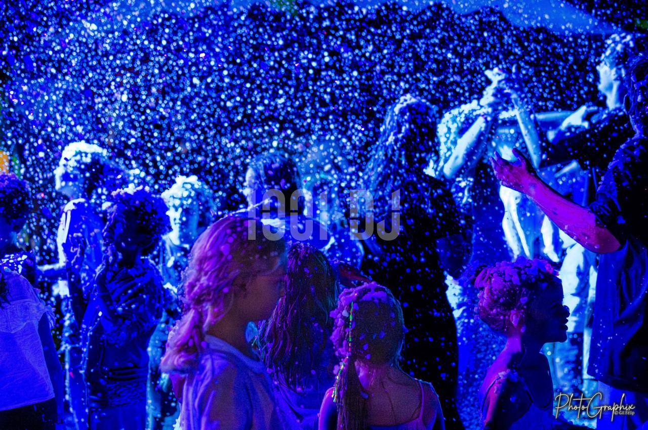 Des enfants et adultes jouent sous une pluie de mousse illuminée par des lumières bleues dans une ambiance festive.