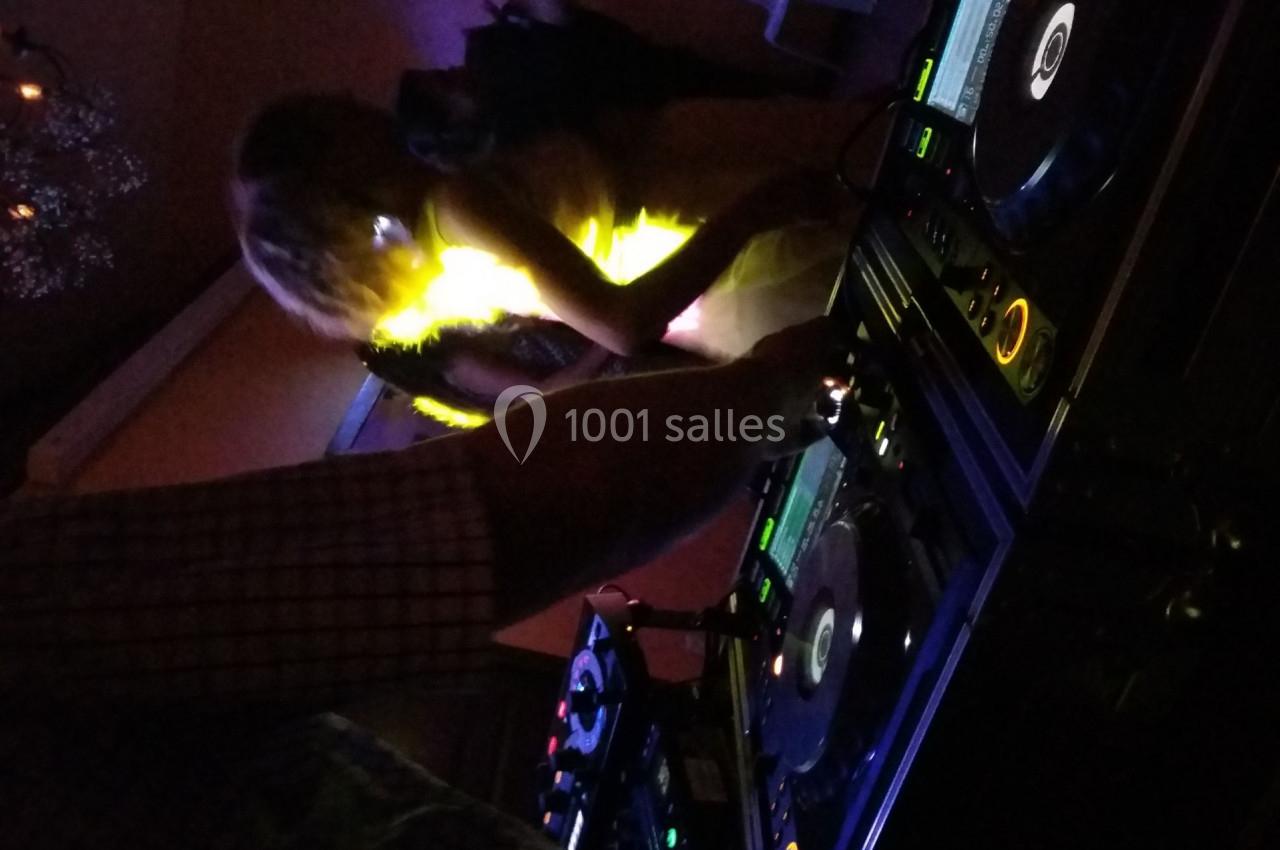 Un DJ mixe sur des platines dans une ambiance sombre avec une personne dansante éclairée par une lumière vive.