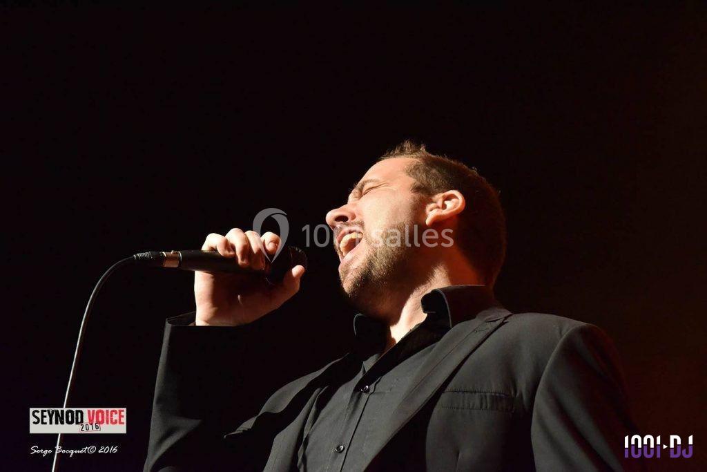 Un homme en costume noir chante dans un micro sur une scène, éclairé par une lumière chaude.