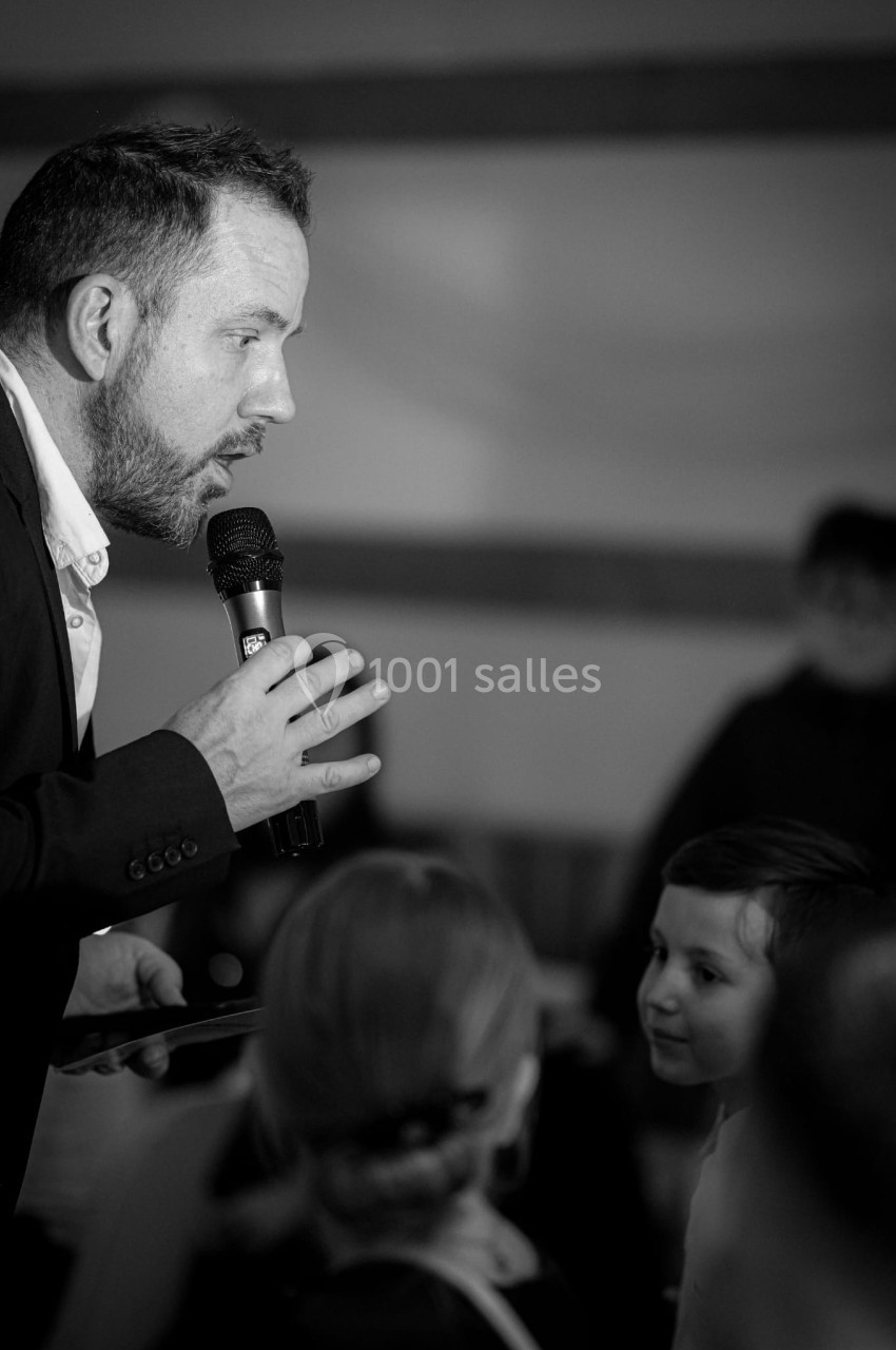 Homme tenant un micro, s'adressant à un enfant dans un cadre intérieur, avec des personnes en arrière-plan.