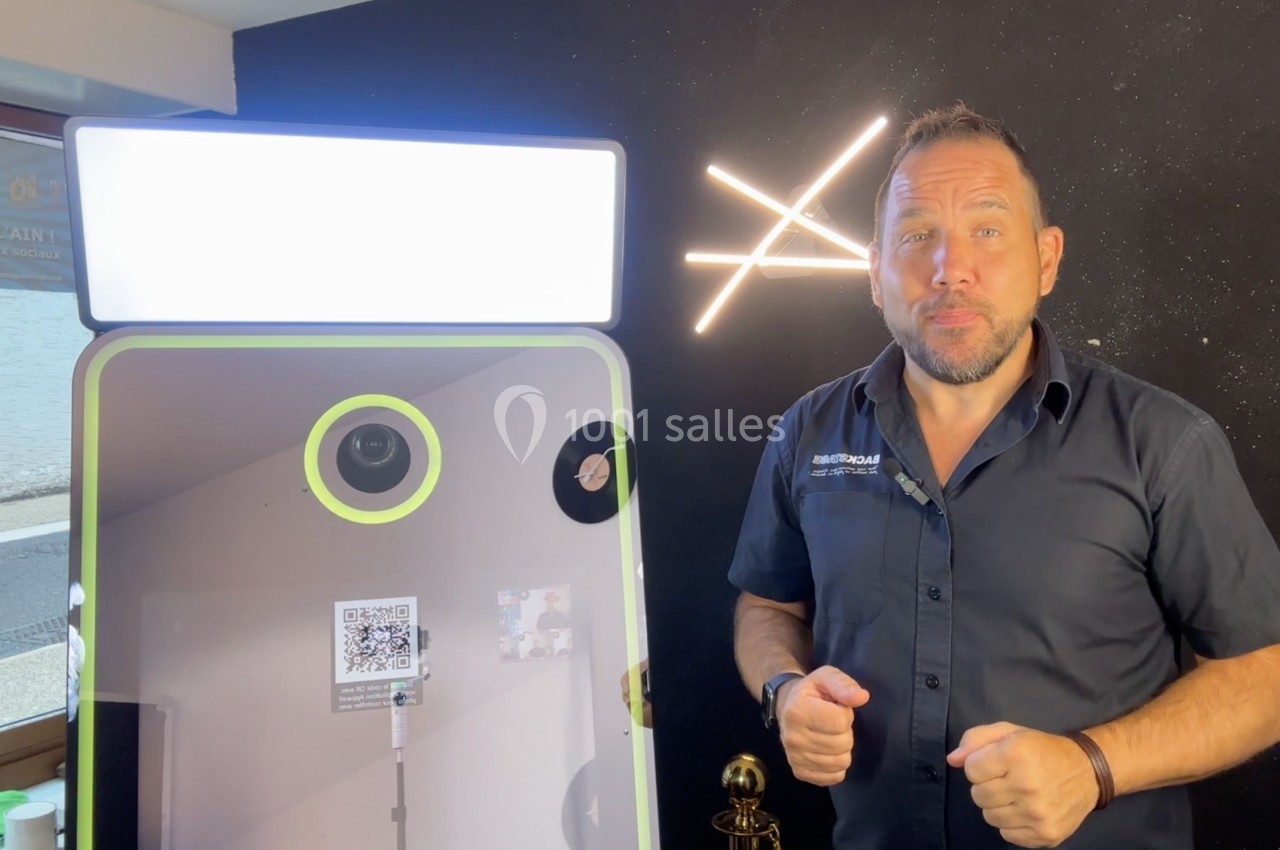 Un homme debout à côté d'une borne photo avec écran lumineux et décor mural sombre en arrière-plan.