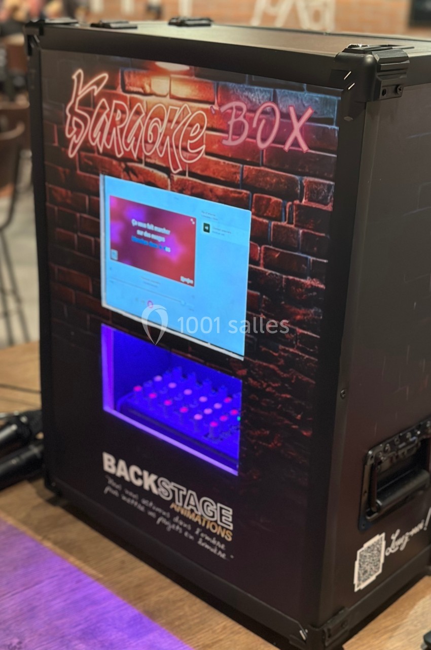 Machine de karaoké noire avec écran tactile, éclairage violet et inscription ’Karaoke Box’ sur un fond de briques rouges.