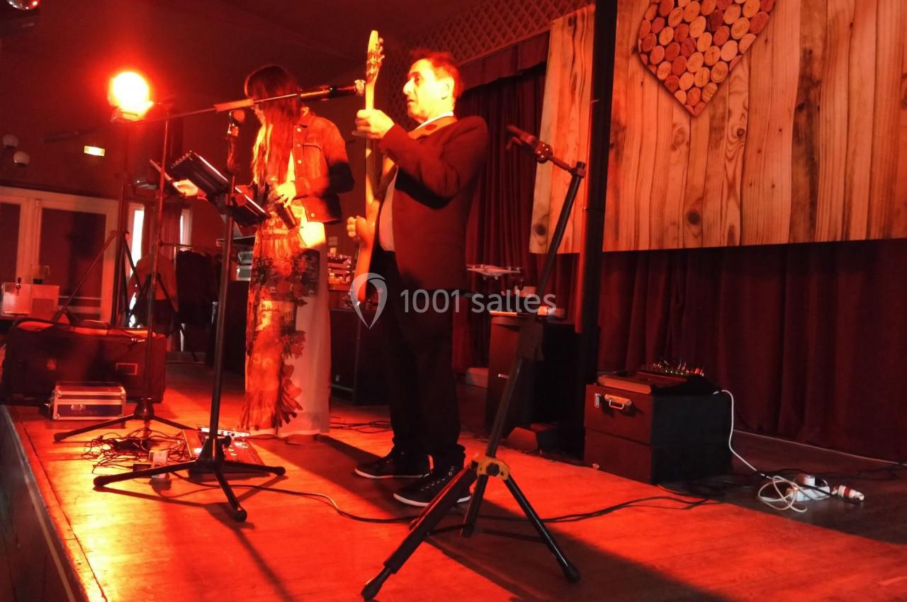 Un duo de musiciens joue sur une scène éclairée en rouge, avec un décor en bois et un cœur en rondins en arrière-plan.